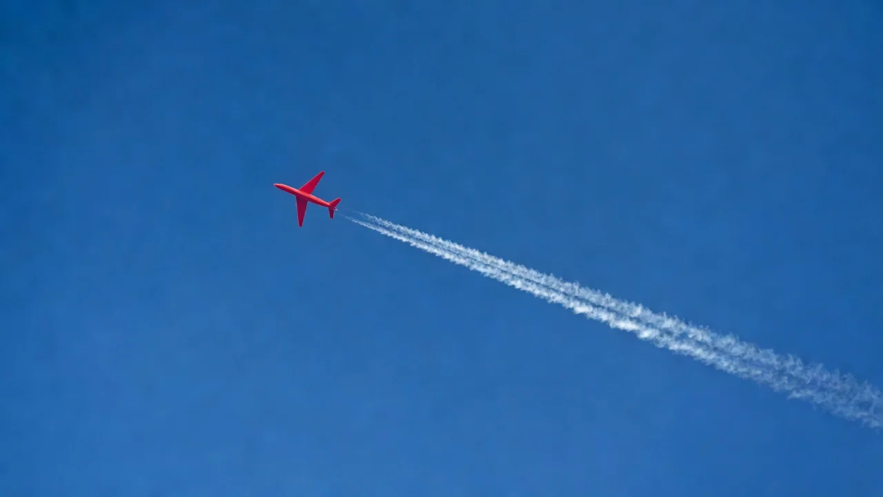 A red plane silhouette against a clear blue sky with a single contrail