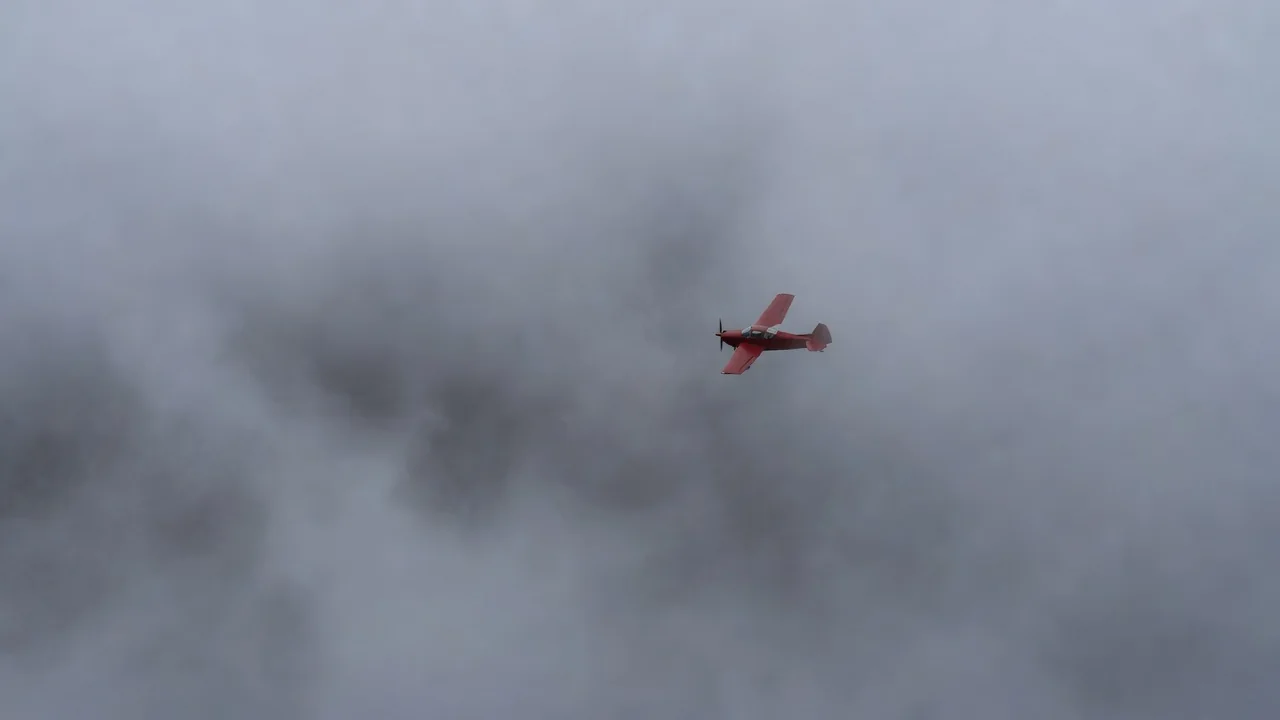 A small red plane flying into a dense fog bank